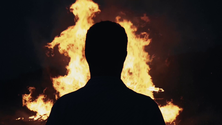 Silhouetted figure stands facing a large bonfire whose tall flames flicker against a darkened backdrop.