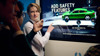 A student in the Road to Zero Experience Space pointing at a touch screen with an image of a green car on the screen 
