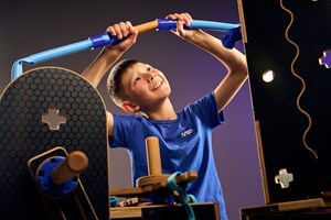 A boy is connecting blue silicone tubes together with a wooden dowel framed by black plywood surfaces dotted with round and cross shaped holes in Tinkertown exhibition at Scienceworks.