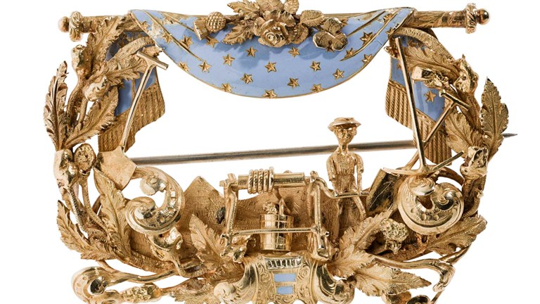 Large gold brooch with openwork foliate design featuring a miner at a windlass, flanked by a pick and shovel on each side of the brooch. The top of the brooch features a blue enameled curtain draped over a gold rod.