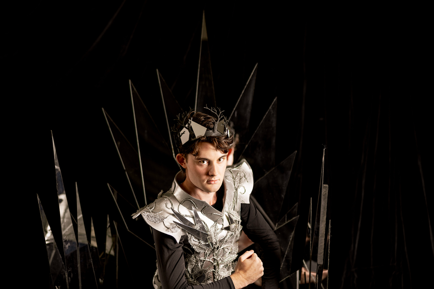 Person wearing a silver and black costume with angular details and a metallic crown, sitting on a throne with sharp, reflective spikes in a dark setting.