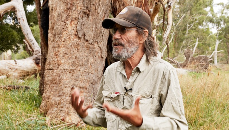Image of older man with a beard and wearing a cap outdoors in front of tree gesticulating while speaking