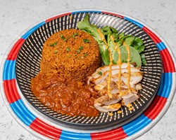 A plate of red rice, grilled chicken, tomato-based curry and green vegetables drizzled with a creamy orange dressing.