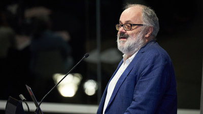 Older man with grey hair, beard and glasses delivering a presentation in a lecture theatre. 