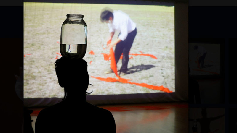  A silhouette of Chung Ngueyn balancing a half full glass jar on their head in front of a video where the artist in a white shirt and black trousers is laying red scarves on the ground.