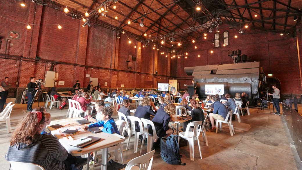 Inside a large brick building are many table and chairs. Students in uniform sit at the tables. Large metal boilers are in the background and in front of them are screens and a speaker sharing slides