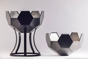 Geodesic metal sphere with hexagonal panels in dark tones, split horizontally in two. The bottom half rests on a curved black-metal stand while the top half sits alongside. 