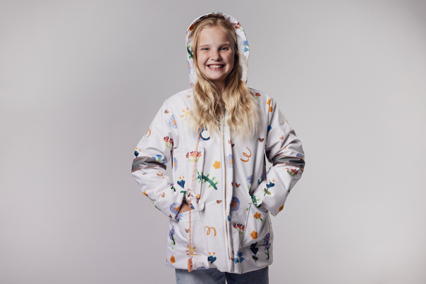 Child wearing a white hooded jacket with colourful patterns and reflective strips smiles with hands in pockets.