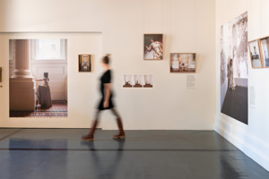 A woman dressed in black is blurred as she quickly moves across a white wall with portraits hanging on it.  