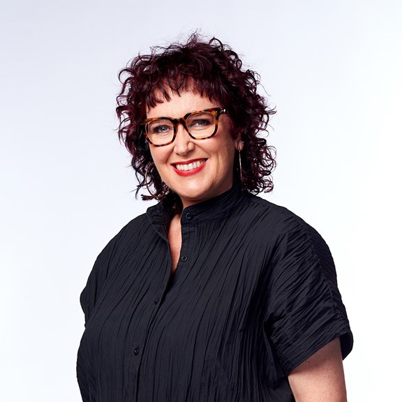 A studio shot of woman with dark curly hair. She is wearing glasses, a black top and bright lipstick.