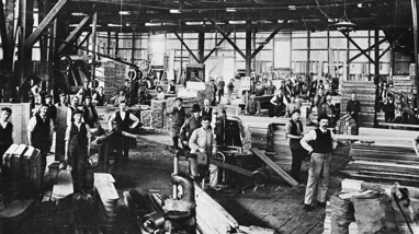 The wood mill in the main factory area. The timber is stacked for manufacturing. Male factory workers stand at the timber and machinery.  