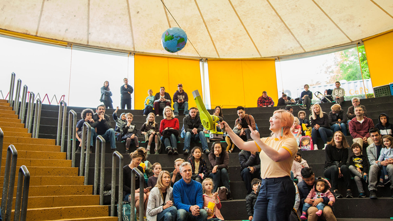 A crowd of people, adults and children, are sitting in a tiered amphitheatre. They are watching a presenter make a small beach ball that looks like the Earth float in the air. The presenter is holding a leaf blower directed at the ball and pointing to the ball with their other hand.