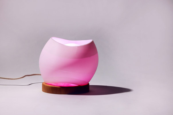 Pink, illuminated lamp with rounded, organic form surrounding a cream-coloured central dome, mounted on a wooden disc.