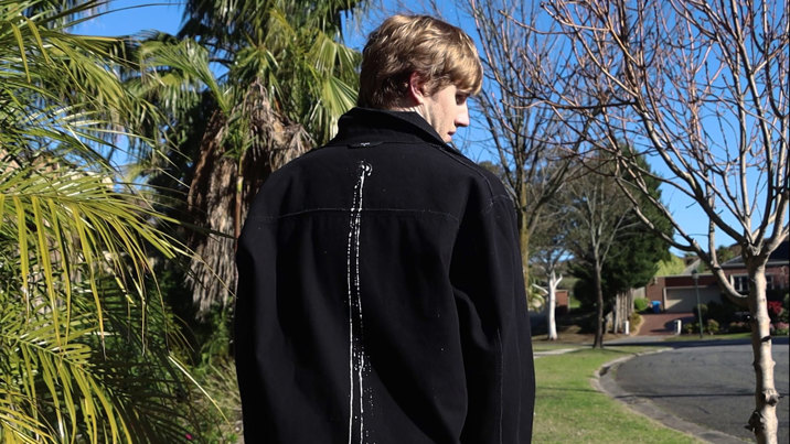 Film still showing a young man in a black jacket walking on a suburban street on a clear day, viewed from behind.