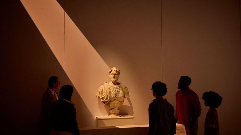 Group of visitors standing around a marble Roman bust on a plinth under dramatic overhead lighting. 