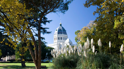The Royal Exhibition Building exterior view on a blue-sky day. The building peaks over trees showing their autumn colours and is reflected in a pond. 
