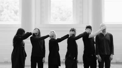 Group of performers standing in a line, each covering the eyes of the person beside them in a grand room.