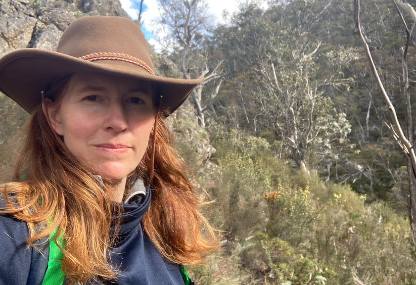 A woman in a brown hat with a large crop of rocks behind her and bushy scrub all around. 