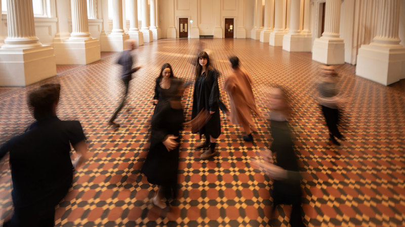 Blurred performers moving through a grand room while one performer stands still at the centre.