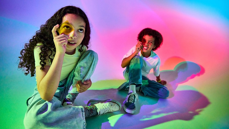 Two young people sit on the floor in a brightly lit room filled with colourful projected lights. The person in the foreground holds up a gold foil packet and a green card toward the camera, while the person behind them sits cross‑legged and smiles. The overlapping pink, blue, purple, and green light patterns create a vibrant, playful atmosphere. 