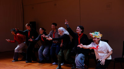 Chung Nguyen in a white shirt, red tie and paper dragon headpiece and interacting with the audience members who all are standing with their hand in a gun-like gesture. 