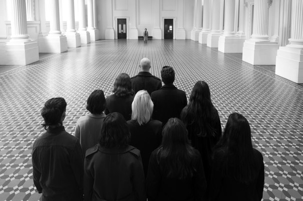  A black and white photo of ten people standing in a wedge formation facing another person on the opposite end of the Long Room at the Immigration Museum.