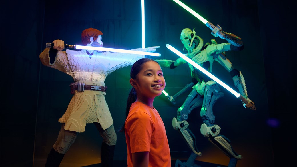 A smiling girl stands in front of a LEGO model of Obi-wan Kenobi fighting General Grievous.
