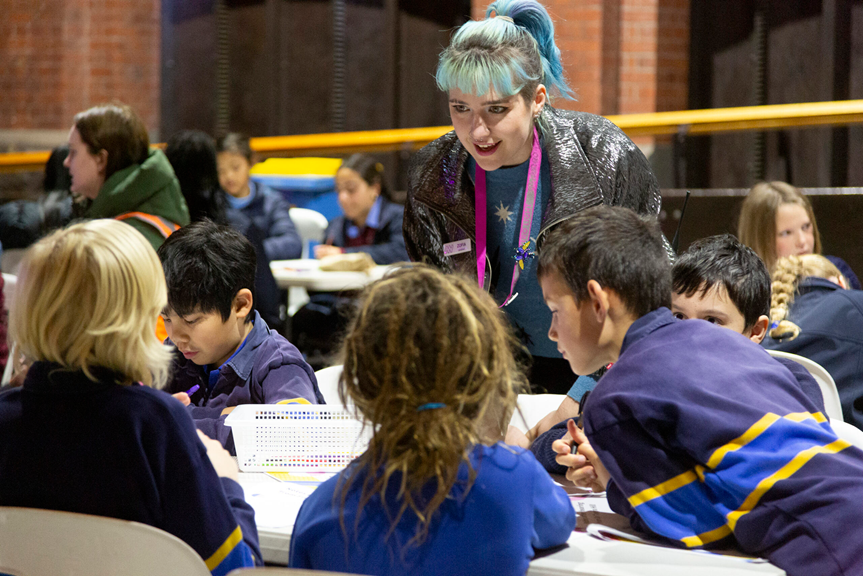 A young adult leans over a table where school students are working. They have pale blue hair and are wearing a pink lanyard that indicates they are a staff member. They look enthusiastic as they talk to the students. 