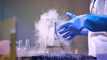 A flask filled with a clear bubbling liquid, a blue rubber glove and the body of a person in a lab coat. The flask has a hazy cloud surrounding it.
