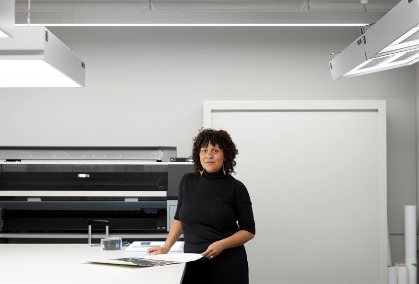 Torika Bolatagici, wearing a black dress, leans against a white table in a large printing room. 