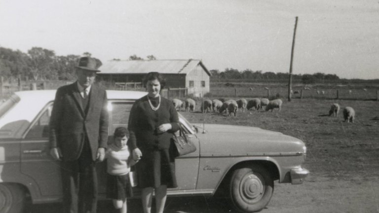 A family of three stand outside a car in front of a farm with sheep grazing in the background.