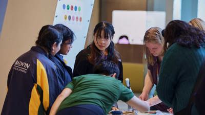 A group of students and a Orygen Youth Mental Health facilitator stand around a table in discussion 