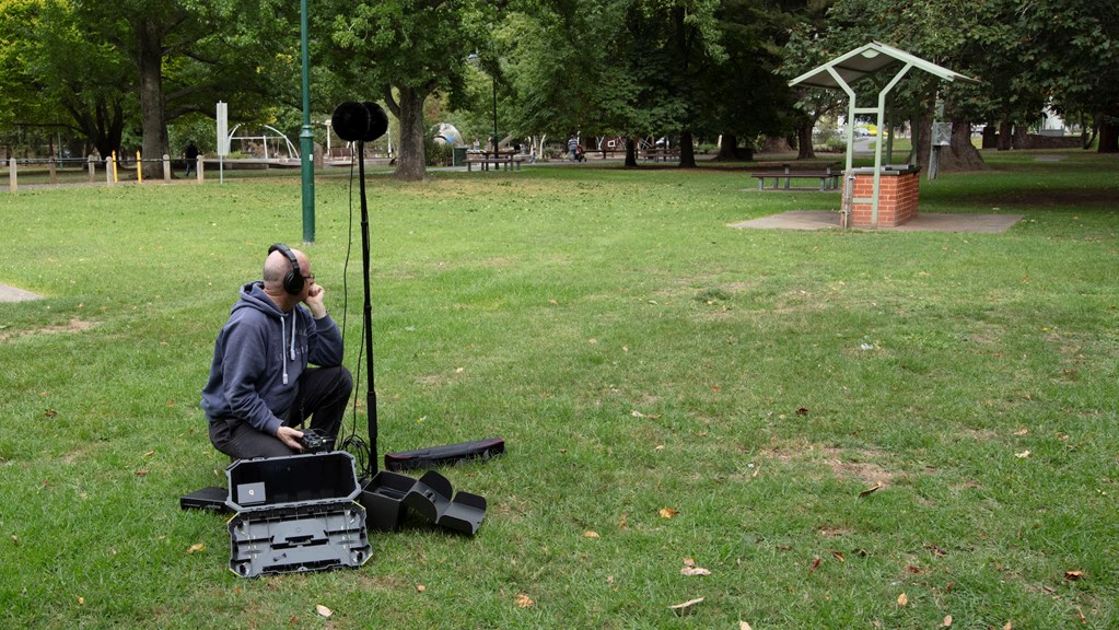 Sound recordist sits on grass besides a large microphone on a stand