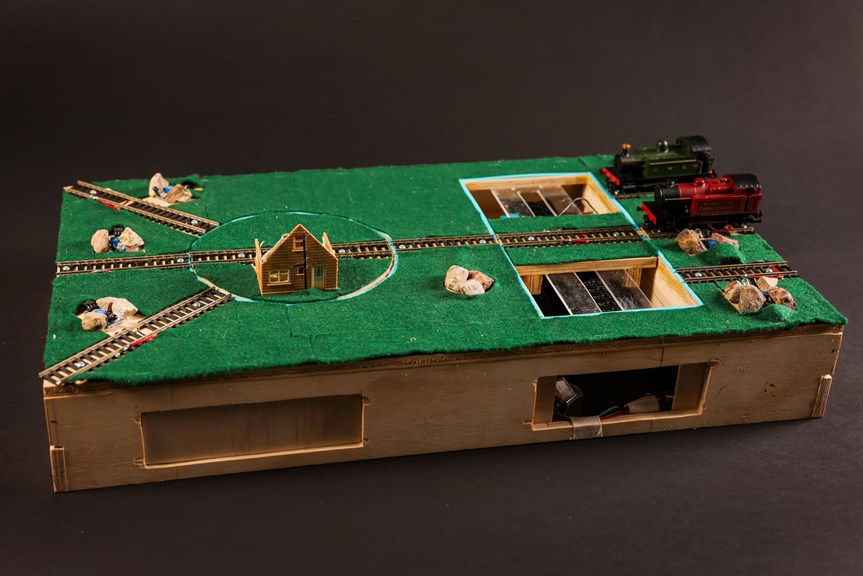 Model trains, train tracks and a small house sit atop a wooden box with a green felt top. System components are visible inside the box.