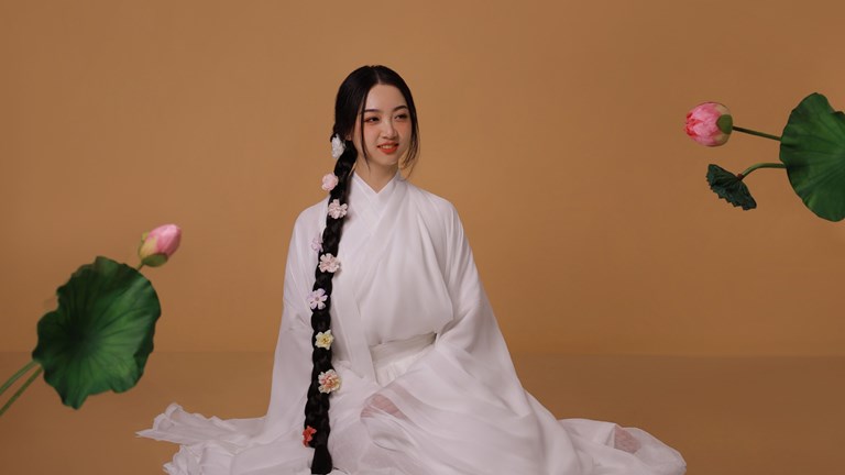 Young person smiling and looking towards the right of frame. They wear a white gown and have long braided hair decorated with flowers.