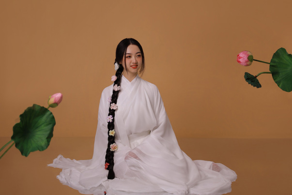 Young person smiling and looking towards the right of frame. They wear a white gown and have long braided hair decorated with flowers.
