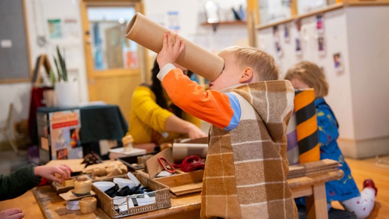 A child in winter warm clothes looks through a cardboard tube like a telescope.