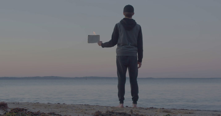 Film still showing a person wearing a hoodie, standing at the water's edge and holding a sign, viewed from behind, with a calm sea under an overcast sky.