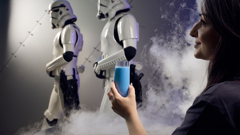 Woman holds blue milk in a smokey room with two stormtroopers in the background.