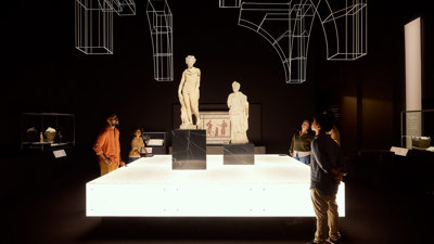 Visitors gathered around two Roman statues on raised plinths within a dramatic, dark exhibition space. 