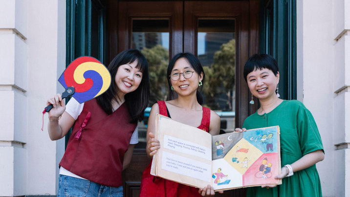 Dr Julie Choi, Youbi Lee and Viet-My Bui are posing with a book in front of the Immigration Museum. 
