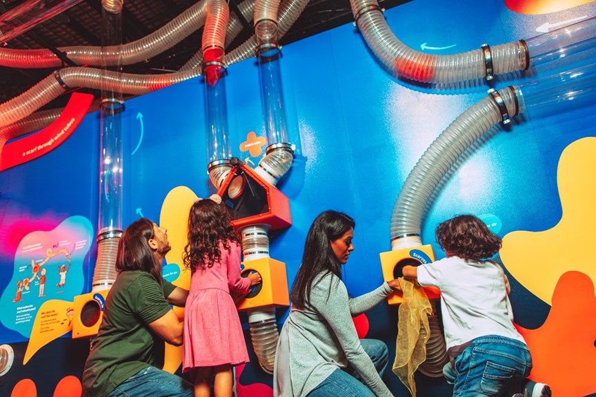 A family playing a game in the Air Playground exhibition. The game is made up of pipes which are mounted on a coloured wall.