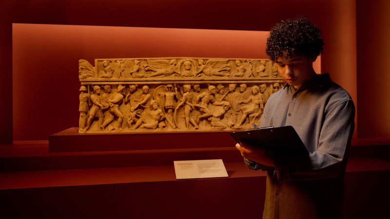 A child writing on a clipboard in front of a roman sarcophagus in ROME: Empire, People Power People Exhibition at Melbourne Museum