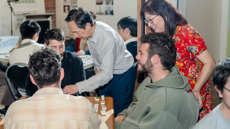 Three young men sit at a square wooden table with Mahjong tiles. Two elderly teachers, one man, and one woman in a red traditional Chinese dress, lean over and teach them how to play. In the background is another table with more players. 