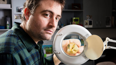 Researcher using an illuminated magnifying lens to closely examine a small fossil or specimen in a laboratory setting.