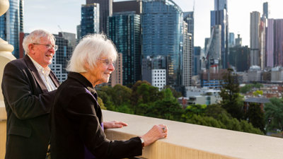 An elderly couple standing on the dome promenade of the Royal exhibition building looking over Carlton Gardens and the Melbourne city skyline.