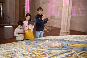 Three children use digital tablets to explore a large, illustrated map of Paris, with a 3D model of the Notre-Dame cathedral in its centre.