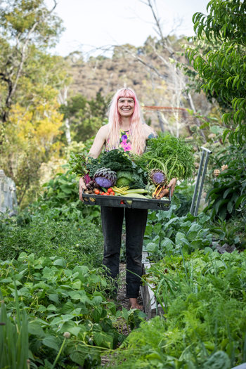 Woman standing in a lush vegetable garden holding a tray of freshly picked vegetables