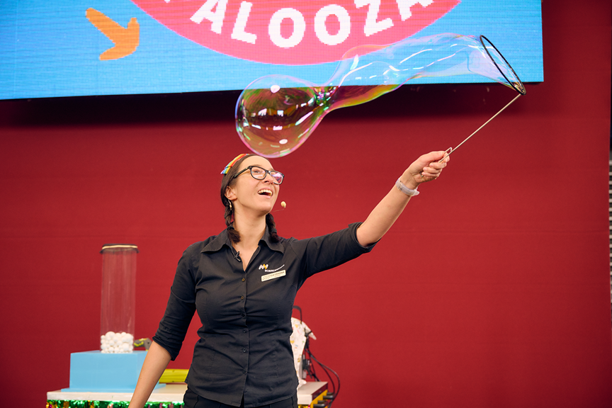 A presenter in a black Scienceworks uniform is holding a large bubble wand and moving it through the air to create a long rainbow bubble. 