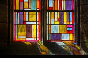 Sunlight shines through the multi-coloured glass panels of a window set in a stone wall.